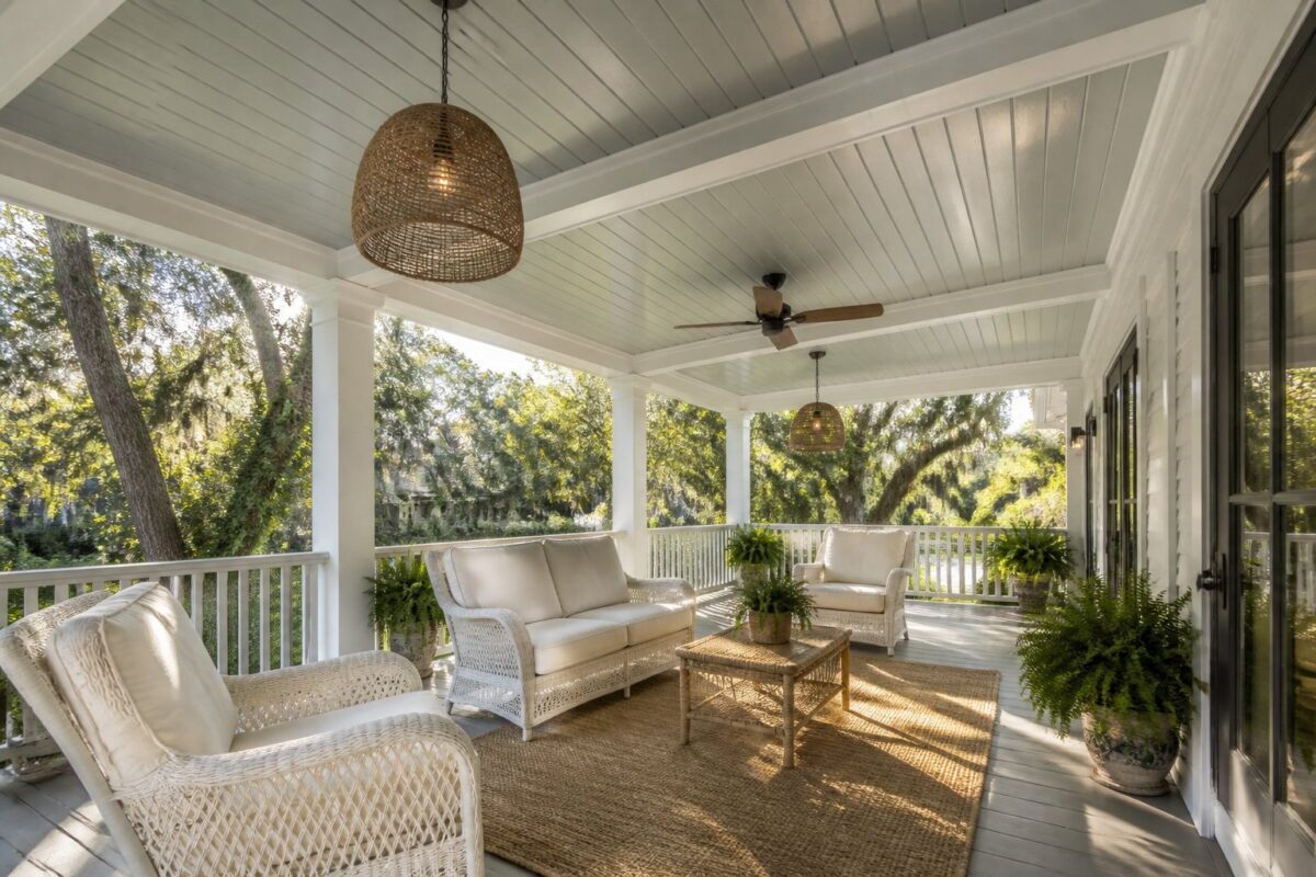 A covered porch with a muted gray-green tongue-and-groove ceiling, white beams, wicker chairs, hanging lanterns, a ceiling fan, potted ferns, and warm sunlight.